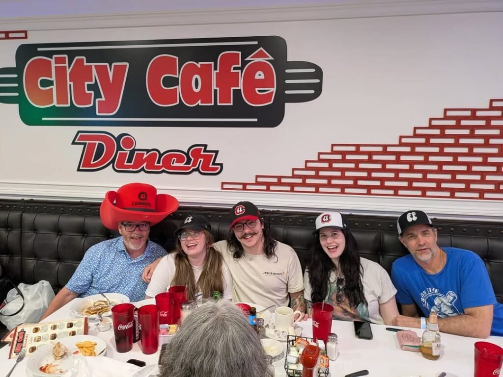Group of five people enjoying a meal together at City Cafe Diner, with plates of food and drinks on the table, smiling and wearing hats, in a cozy diner setting.